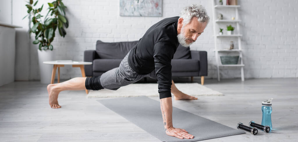 hombre descalzo con el pelo gris haciendo ejercicio en la estera de fitness cerca de pesas en la sala de estar, pancarta - Foto, Imagen