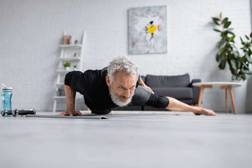 silny mężczyzna z siwymi włosami ćwiczący na macie fitness w pobliżu hantli w salonie - Zdjęcie, obraz