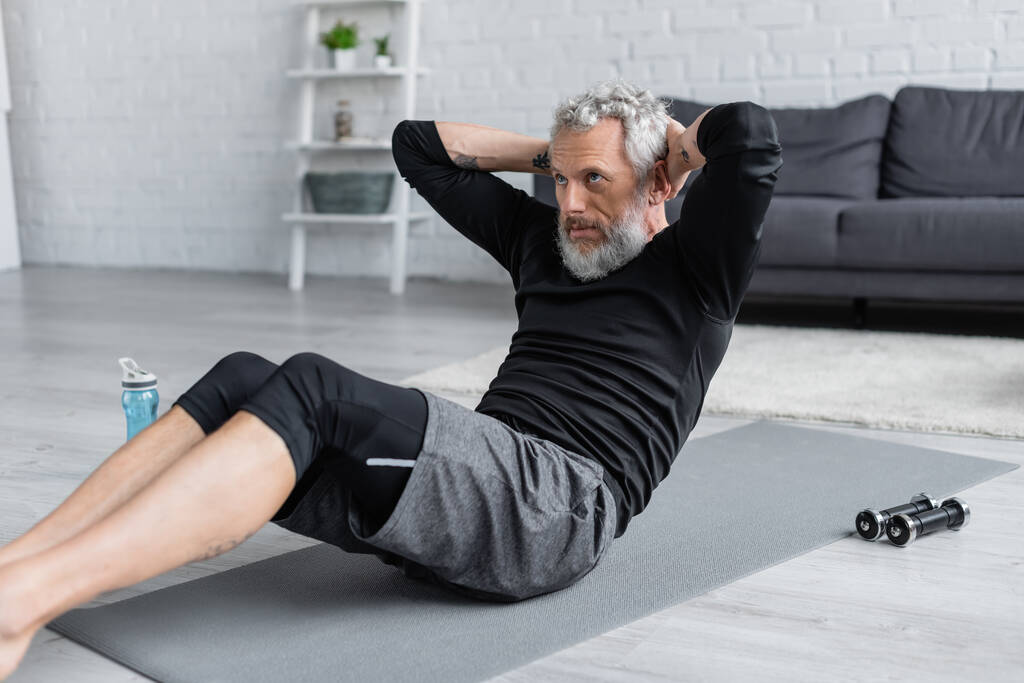 bärtiger und tätowierter Mann trainiert auf Fitnessmatte im Wohnzimmer - Foto, Bild