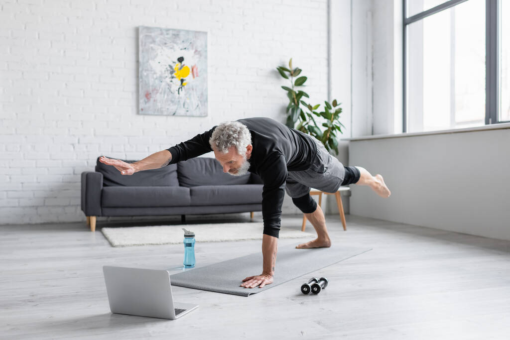 homem com cabelos grisalhos exercitando no tapete de fitness perto de laptop na sala de estar  - Foto, Imagem