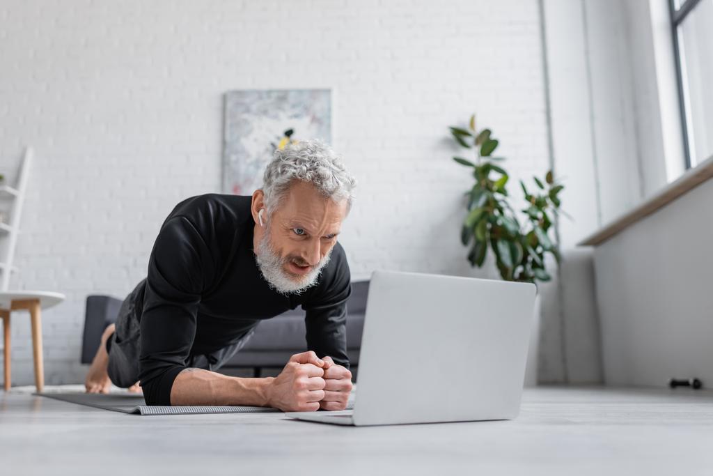 Kablosuz kulaklıklı sakallı adam oturma odasındaki dizüstü bilgisayarın yanında spor tahtası yapıyor.  - Fotoğraf, Görsel