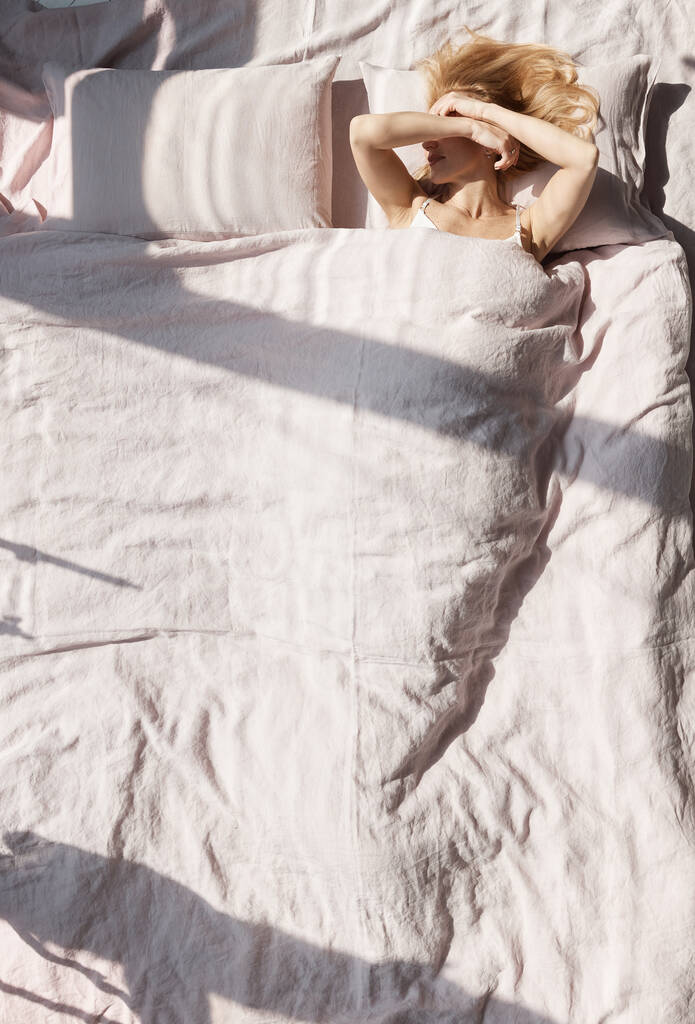 kép egy gyönyörű szőke nő feküdt egy vászon pasztell ágynemű eltakarja arcát a nap, amely megvilágítja a szobát - Fotó, kép