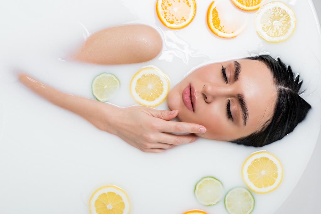 top view of young woman touching face while bathing in milk with citrus slices - Photo, Image