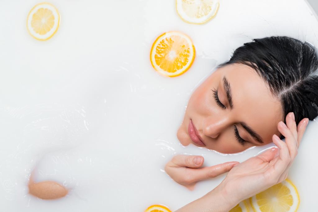 vista superior da mulher bonita com olhos fechados tomando banho de leite com limão e fatias de laranja - Foto, Imagem