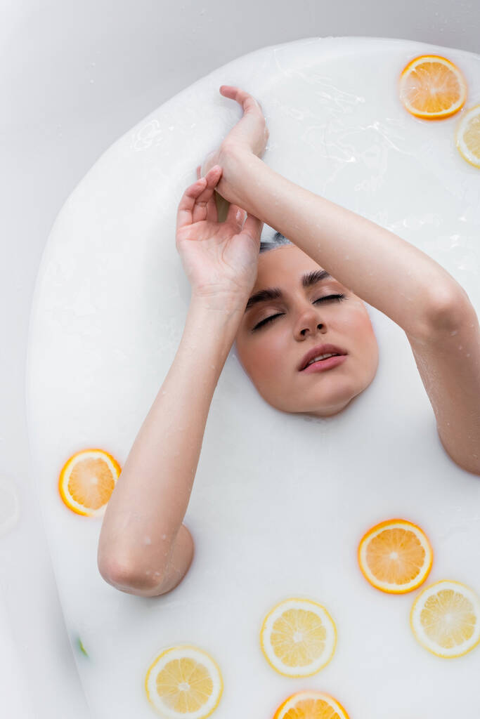 młoda kobieta z zamkniętymi oczami kąpiąca się w mleku ze świeżymi cytrusami - Zdjęcie, obraz
