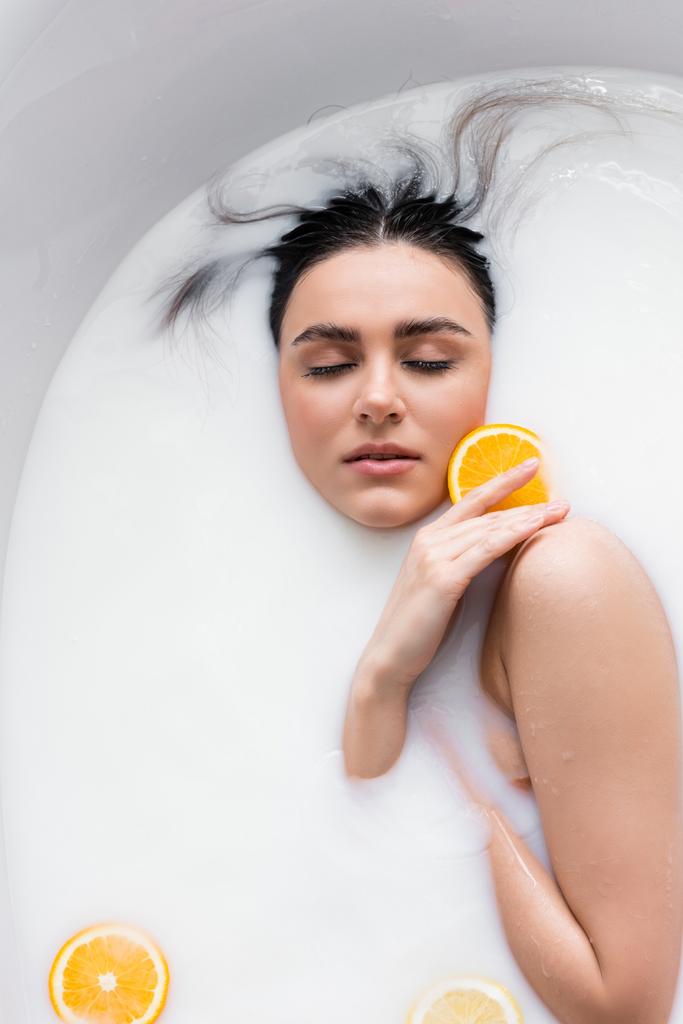 jovem com olhos fechados segurando fatia de laranja enquanto relaxa no banho de leite - Foto, Imagem
