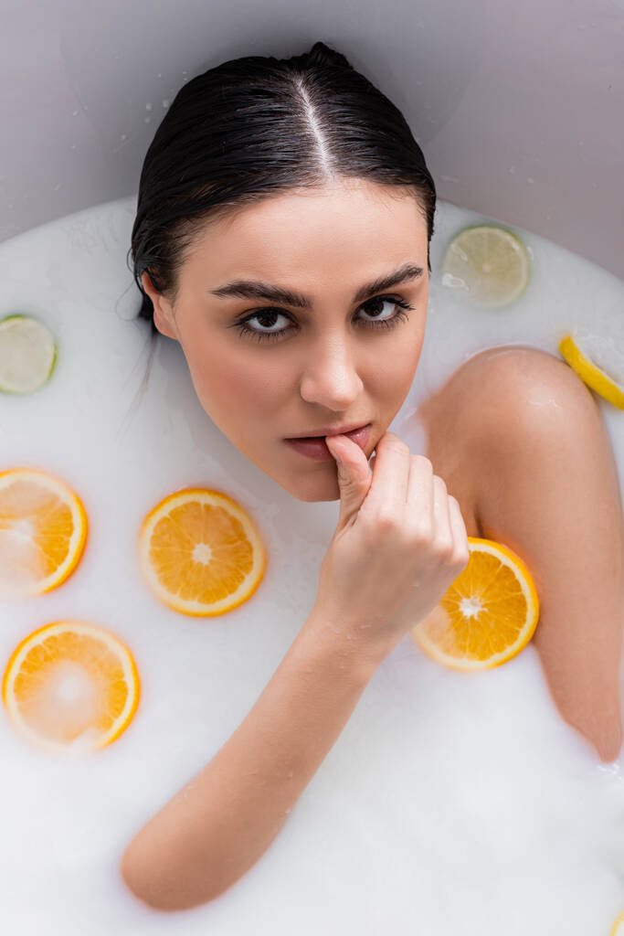 sensuele vrouw in melk bad met citrusvruchten aanraken lippen terwijl kijken naar camera - Foto, afbeelding