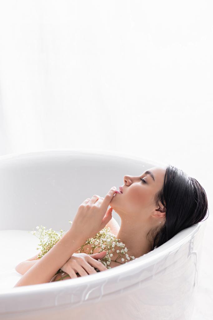 giovane donna toccando mento e tenendo fiori di gypsophila durante il bagno nel latte - Foto, immagini