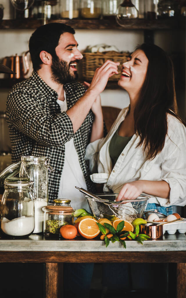 Mladý šťastný pár vaří spolu v kuchyni. Pozitivní běloch a bruneta si hrají s moukou, flirtují, smějí se a míchají přísady do těsta na pečení citrusového dortu. Valentýn - Fotografie, Obrázek
