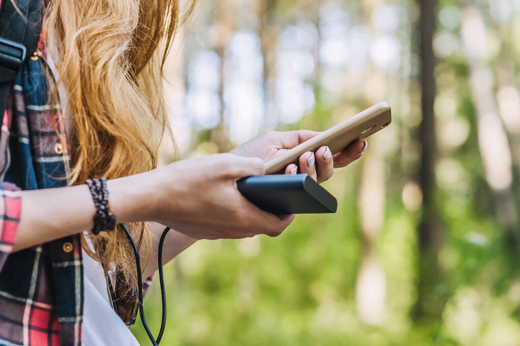 Power banka a smartphone v rukou dívky s rudými vlasy v košili v kleci s černým batohem, na pozadí lesní cesty - Fotografie, Obrázek