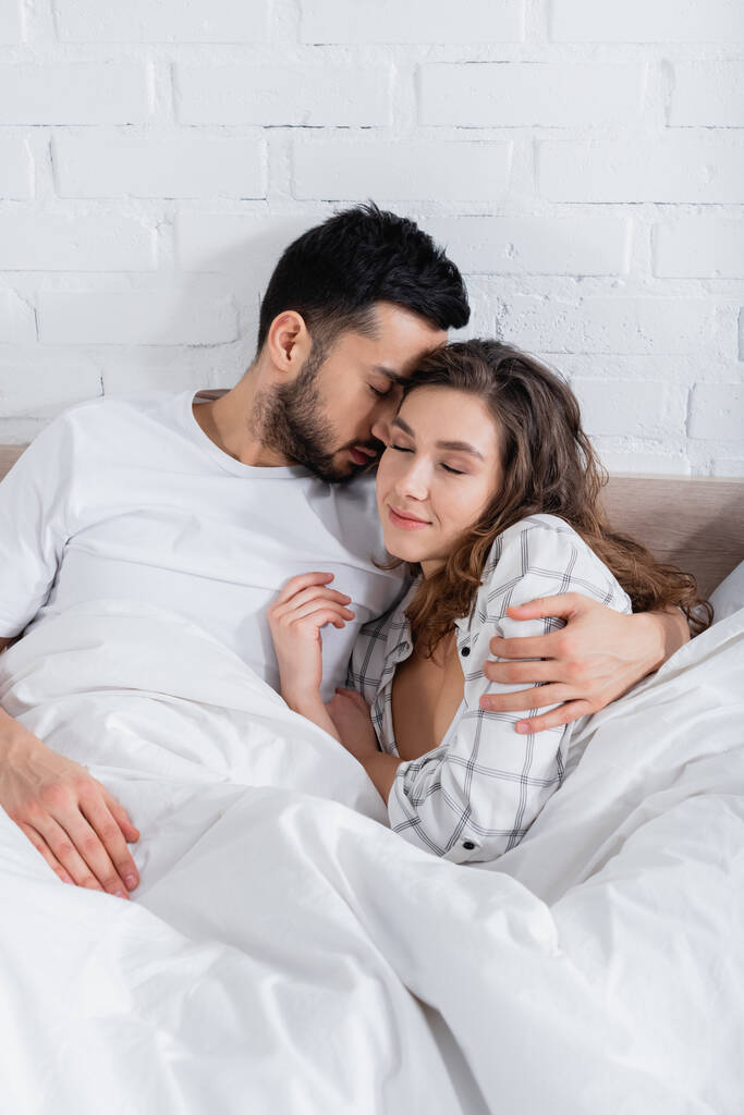 Sakallı Müslüman adam yatakta gözleri kapalı kız arkadaşına sarılıyor. - Fotoğraf, Görsel