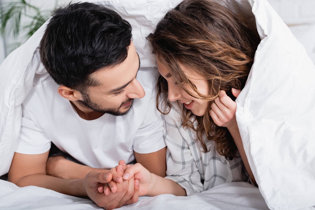 sourire interracial couple tenant la main tout en étant couché sous la couverture  - Photo, image