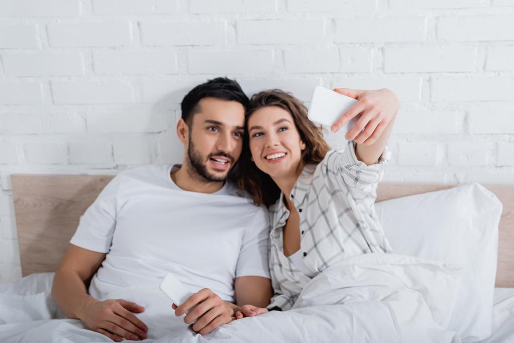 mulher feliz tomando selfie com barbudo muçulmano homem na cama - Foto, Imagem
