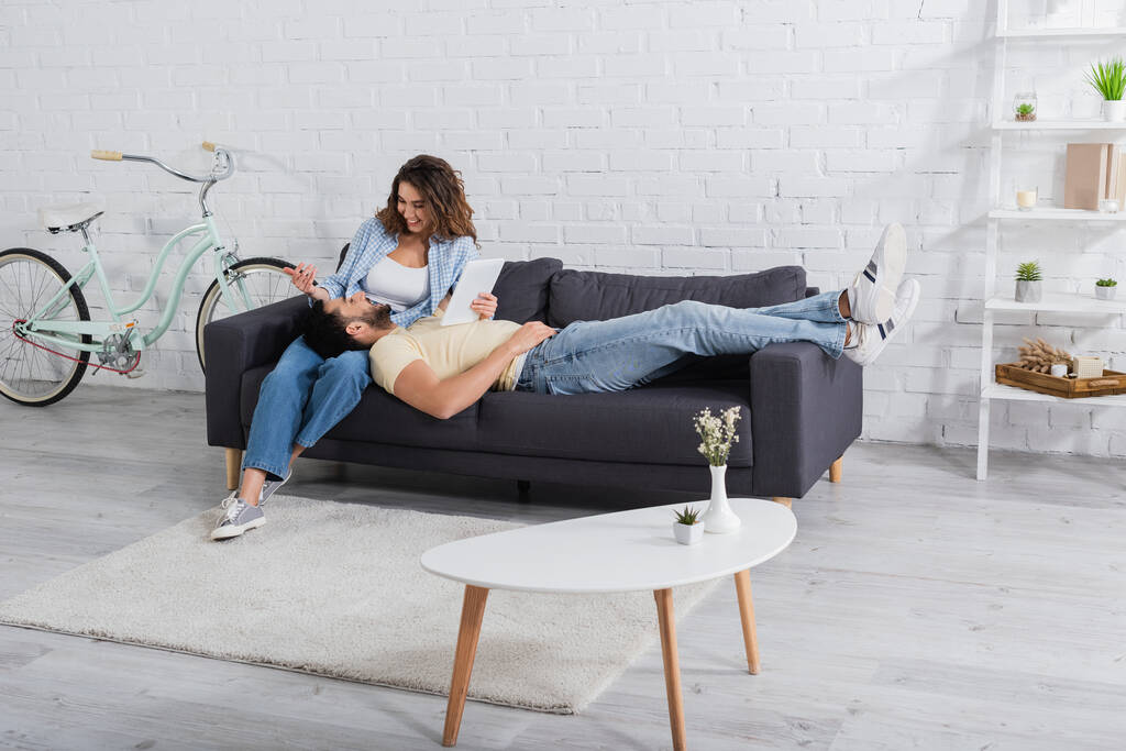 homem muçulmano descansando no sofá com tablet digital perto de namorada feliz  - Foto, Imagem