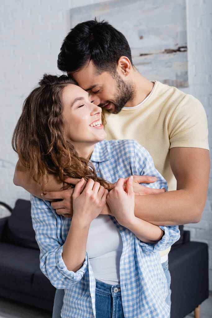 glimlachende multi-etnische man en vrouw met gesloten ogen knuffelen thuis - Foto, afbeelding