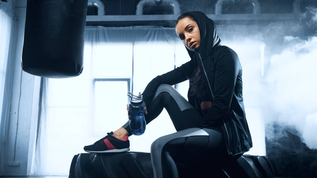 donna sportiva in felpa con cappuccio seduta su pneumatico con bottiglia sportiva e guardando la fotocamera  - Foto, immagini