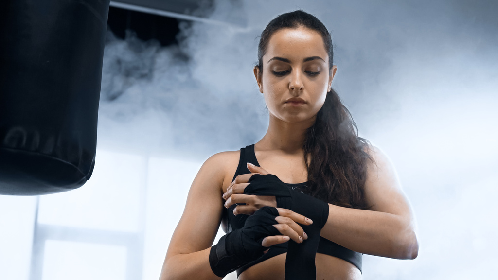 young brunette sportswoman bandaging hands near punching bag  - Photo, Image