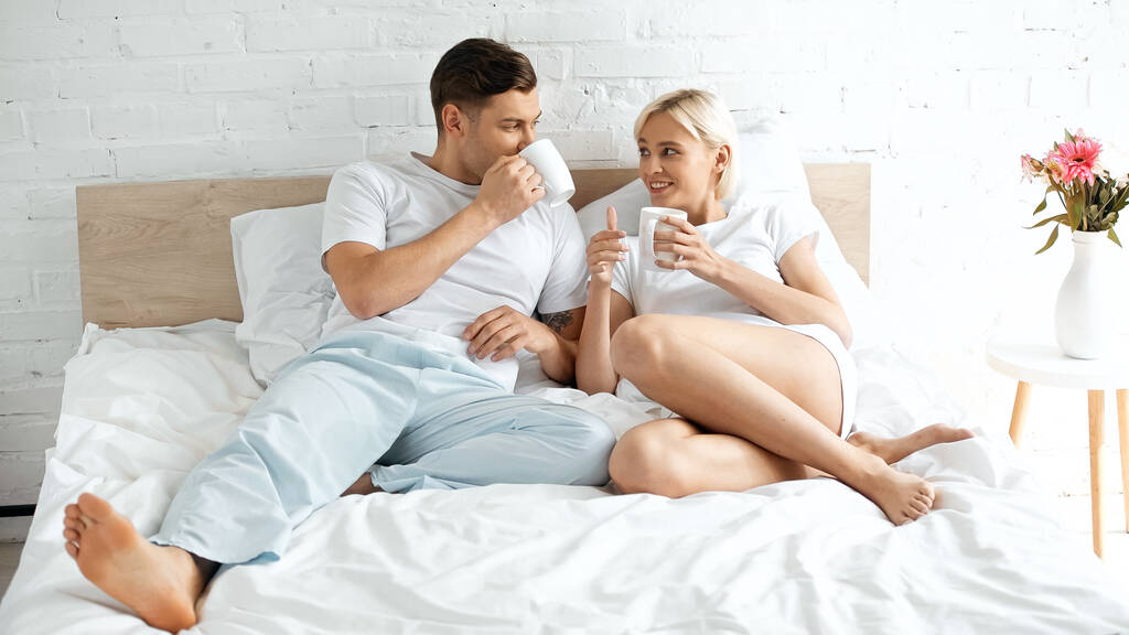uśmiechnięta młoda kobieta patrząc na chłopaka pijącego kawę w łóżku - Zdjęcie, obraz