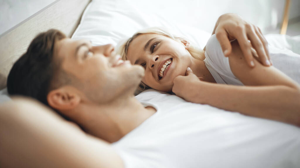 glückliche Frau lächelt, während sie mit Freund auf verschwommenem Vordergrund im Bett liegt  - Foto, Bild