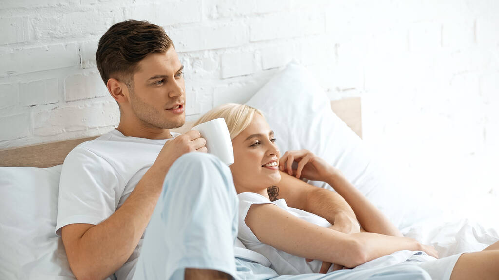 wytatuowany mężczyzna trzymający filiżankę kawy w pobliżu szczęśliwa kobieta leżąca w łóżku  - Zdjęcie, obraz