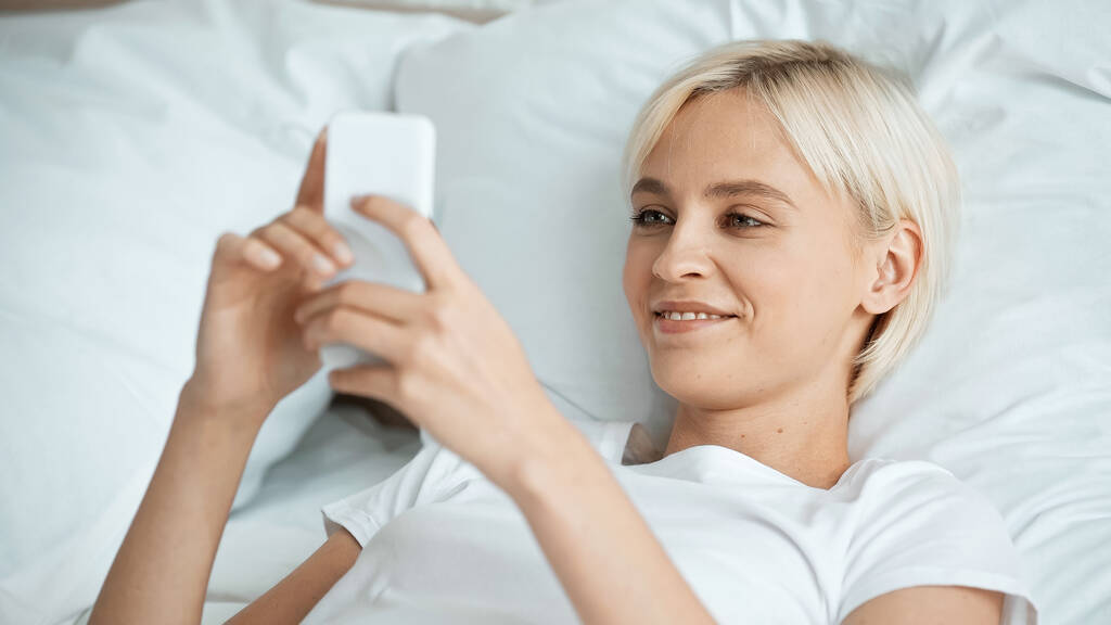 pleased young woman texting on smartphone in bedroom  - Photo, Image