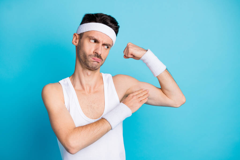 Фото неуверенного спортивного парня смотреть его руки мышцы носить белый цистерны топ синглет изолированы на синем фоне цвета - Фото, изображение