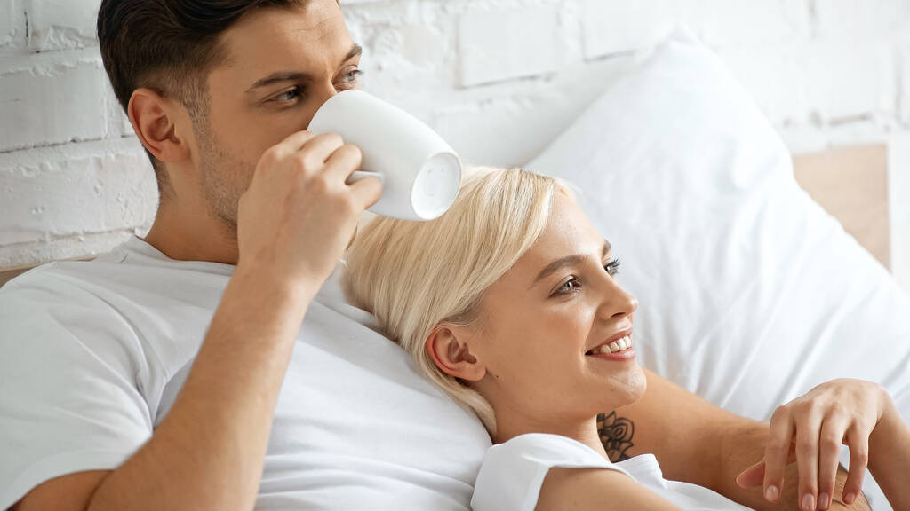 tattooed man drinking coffee while lying with pleased blonde woman in bed - Photo, Image