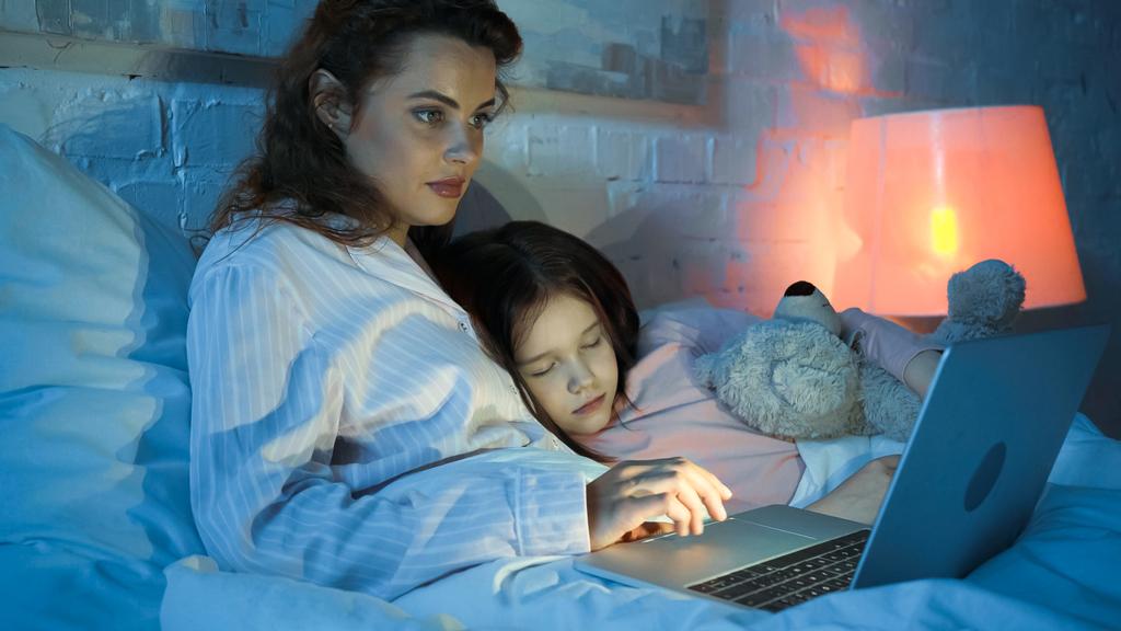 Frau benutzte Laptop neben schlafendem Kind auf Bett  - Foto, Bild