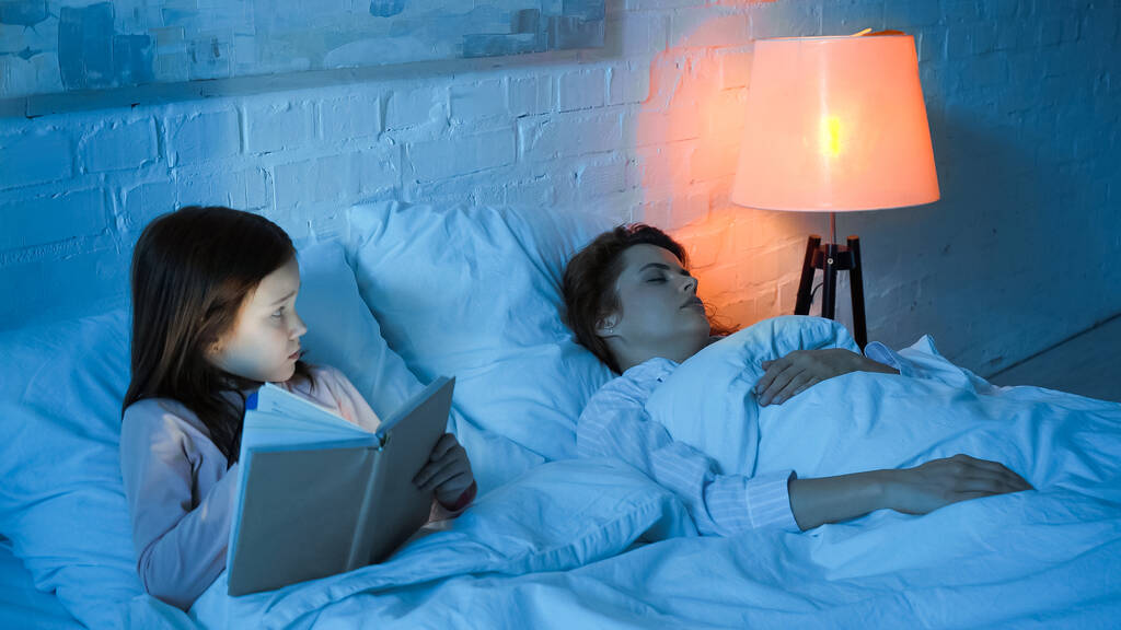 Mädchen mit Buch sieht schlafende Mutter nachts im Bett an  - Foto, Bild