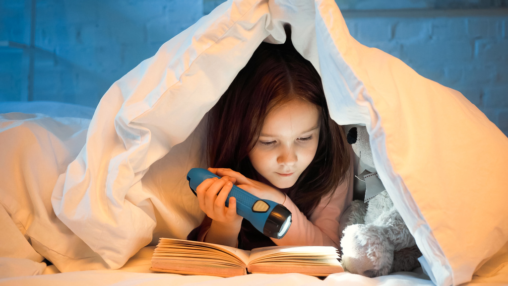 Preteen kid holding flashlight while reading book near soft toy on bed  - Photo, Image