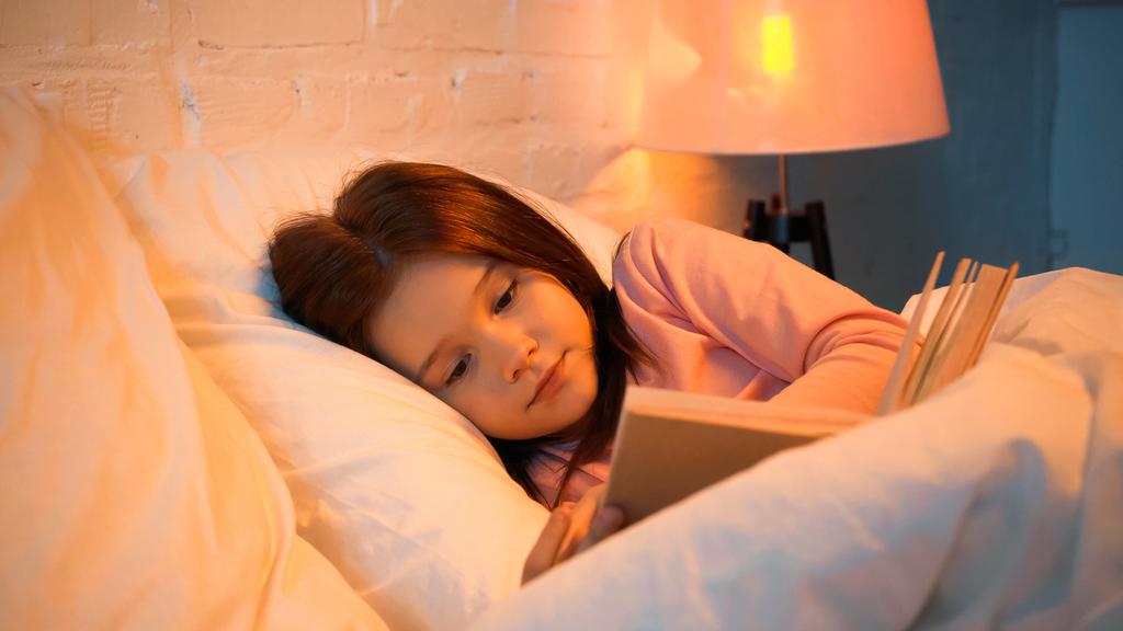 Kid leitura livro perto de cobertor borrado na cama  - Foto, Imagem