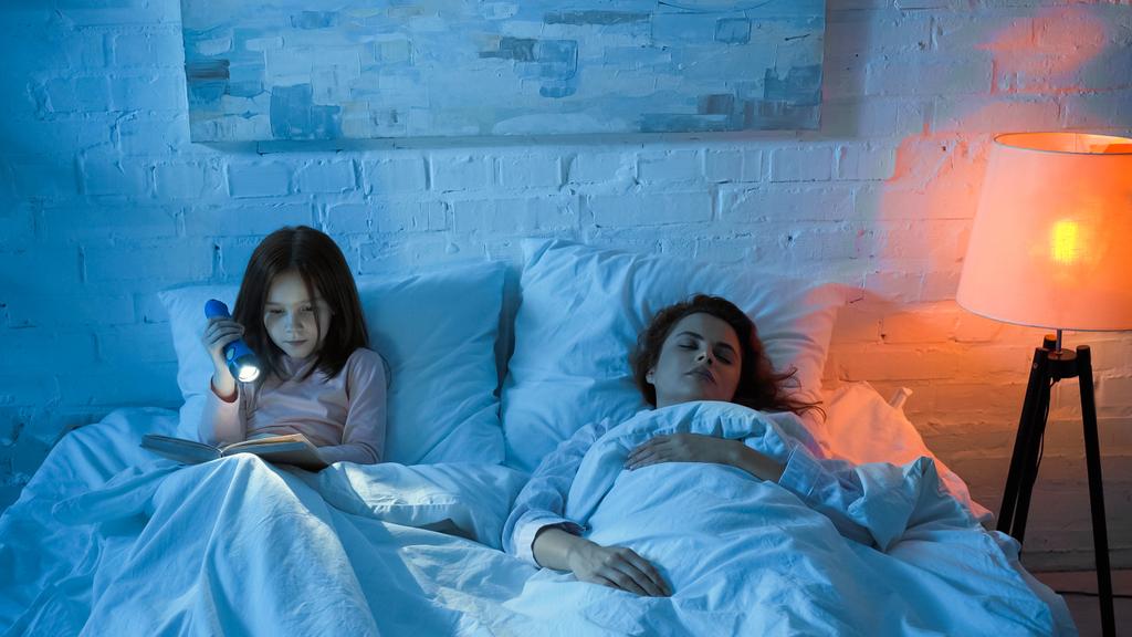 夜にベッドで本を読んでいる子供の近くで寝る女性  - 写真・画像