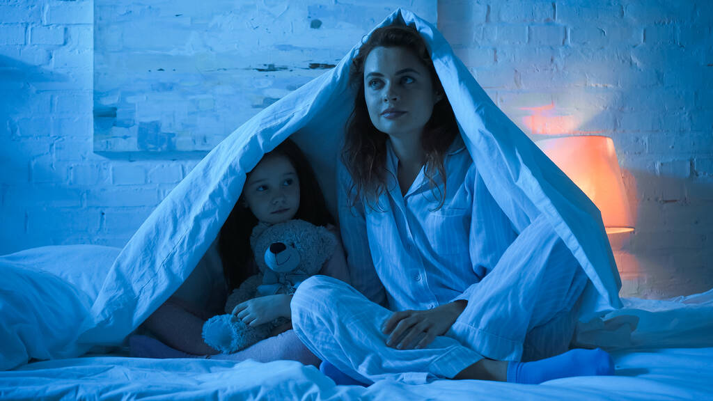 Moeder en kind met zacht speelgoed zitten onder deken op bed tijdens de nacht  - Foto, afbeelding