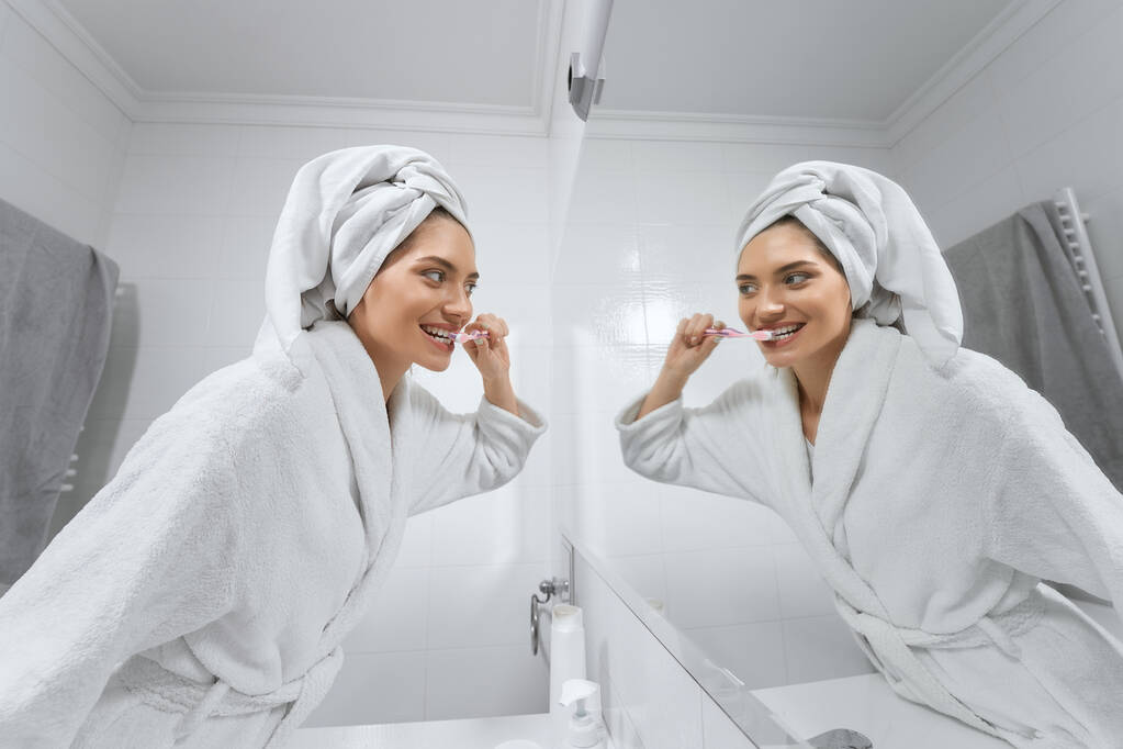 Mujer en bata blanca con toalla en la cabeza cepillándose los dientes.  - Foto, Imagen