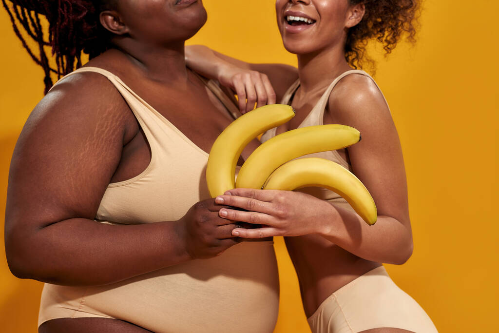 Schnappschuss von plus size und schlanke junge afrikanisch-amerikanische Frauen in Unterwäsche posieren mit reifen gelben Bananen isoliert über orangefarbenem Hintergrund - Foto, Bild