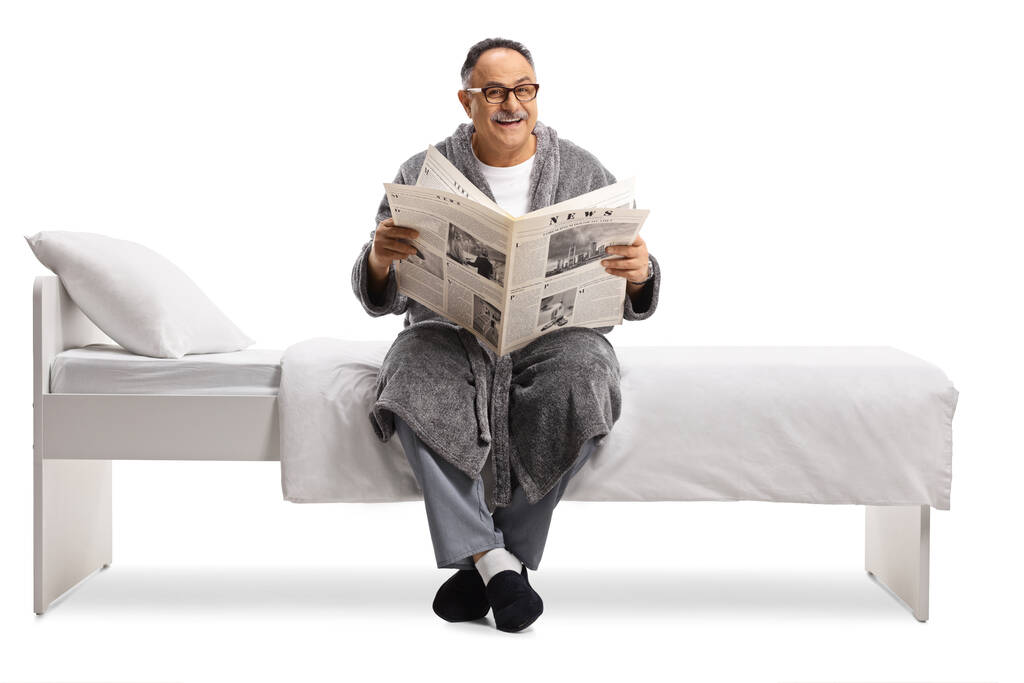 Starší muž v županu sedí na posteli a čte noviny izolované na bílém pozadí - Fotografie, Obrázek