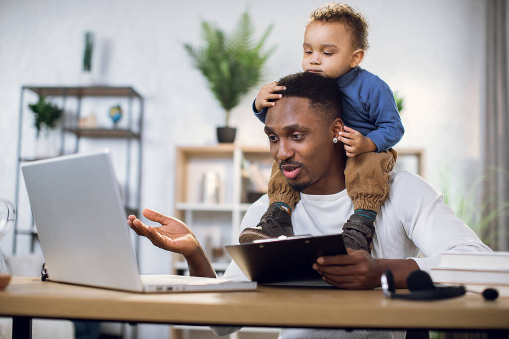 Зайнятий афро людина працює на ноутбуці з сином на шиї
 - Фото, зображення