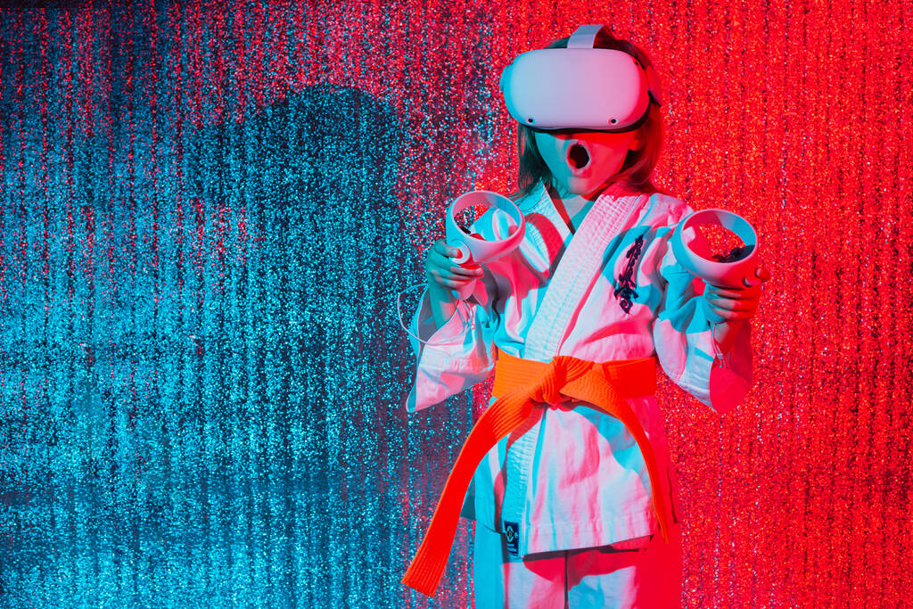 Studioporträt eines jungen kaukasischen Mädchens in Karate-Uniform, das Videospiele mit Virtual-Reality-Headset spielt oder online mit einem Trainer auf blauem und rotem Hintergrund übt. Zukünftiges Technologiekonzept. VR - Foto, Bild