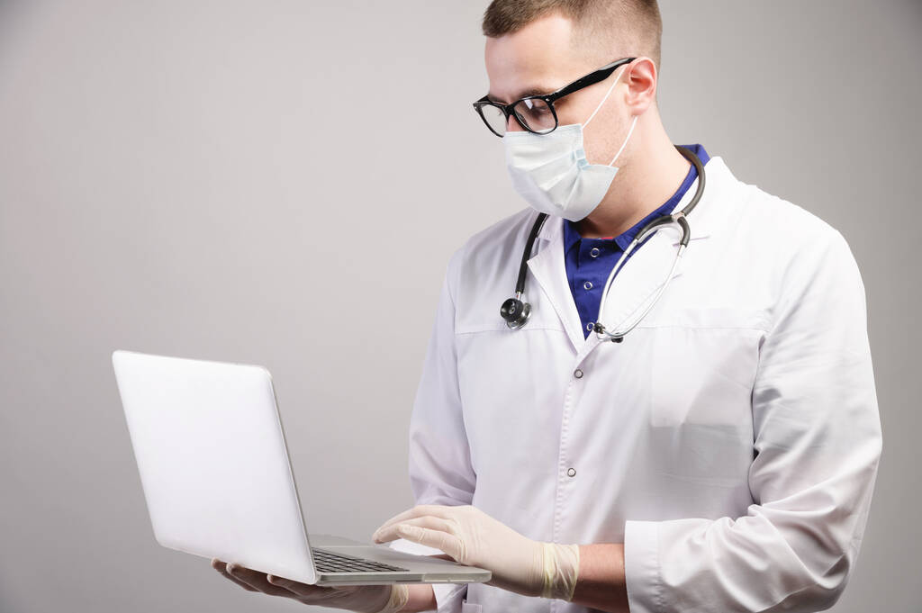 mladý běloch v masce používá svůj notebook pro lékařské účely na šedém pozadí. Studio shot - Fotografie, Obrázek
