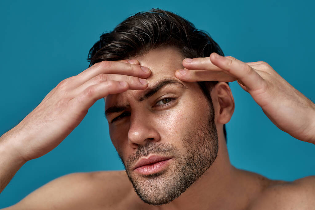 Nahaufnahme Porträt eines hemdlosen, gutaussehenden Typen, der seine Haut überprüft, auf der Suche nach Makeln, während er isoliert vor blauem Hintergrund posiert - Foto, Bild