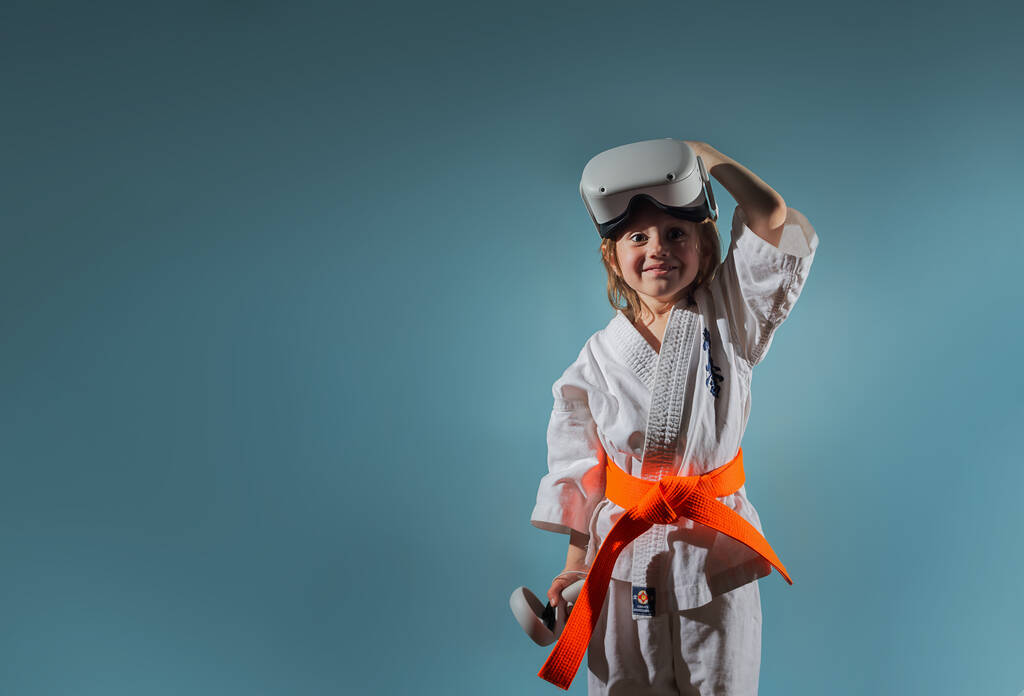 Studio Portrait de jeune fille caucasienne en uniforme de karaté jouant à des jeux vidéo avec casque de réalité virtuelle ou pratiquant en ligne, texte brodé sur kimono kyokushinkai. Bannière. - Photo, image