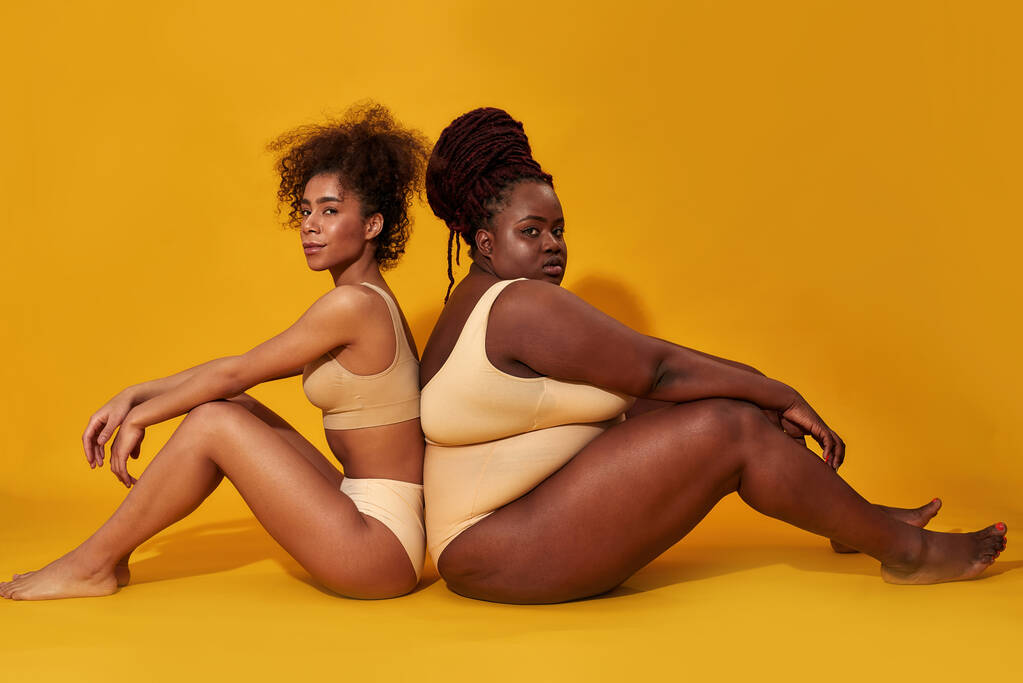 Tiro de comprimento total de dois diversos modelos femininos afro-americanos em roupa interior sentados juntos isolados sobre fundo amarelo - Foto, Imagem