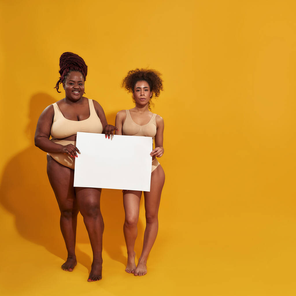 2人のアフリカ系アメリカ人の天然の女性の完全な長さのショット異なる体形状を持つ下着カメラで笑顔,空白のバナーを保持,黄色の背景に隔離された立って - 写真・画像