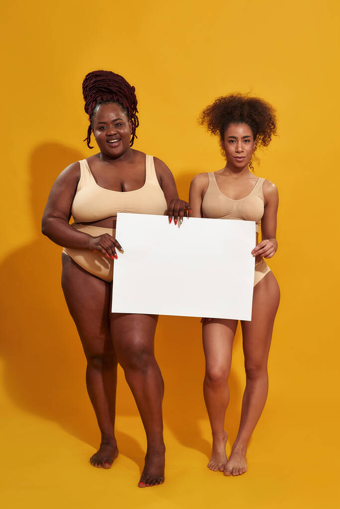 Täyspitkä laukaus kahdesta nuoresta afrikkalaisesta amerikkalaisesta naisesta alusvaatteissa, joiden vartalo on erilainen, katselemassa kameraa, pitämässä tyhjää valkoista lippua, seisomassa eristettynä keltaisen taustan päällä - Valokuva, kuva