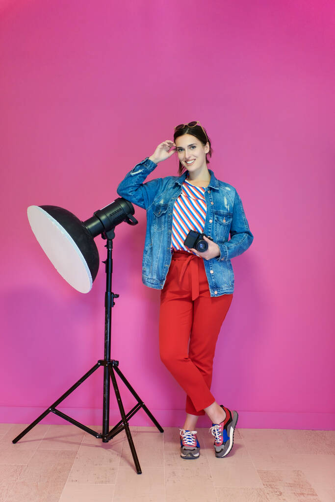 Nuori nainen nojaa studion taskulamppua vasten ja pitelee kameraa vaaleanpunaisella taustalla. Naisvalokuvaaja yllään muodikas ja rento vaatteita poseeraa studiossa. Ammattimainen valokuvaus - Valokuva, kuva