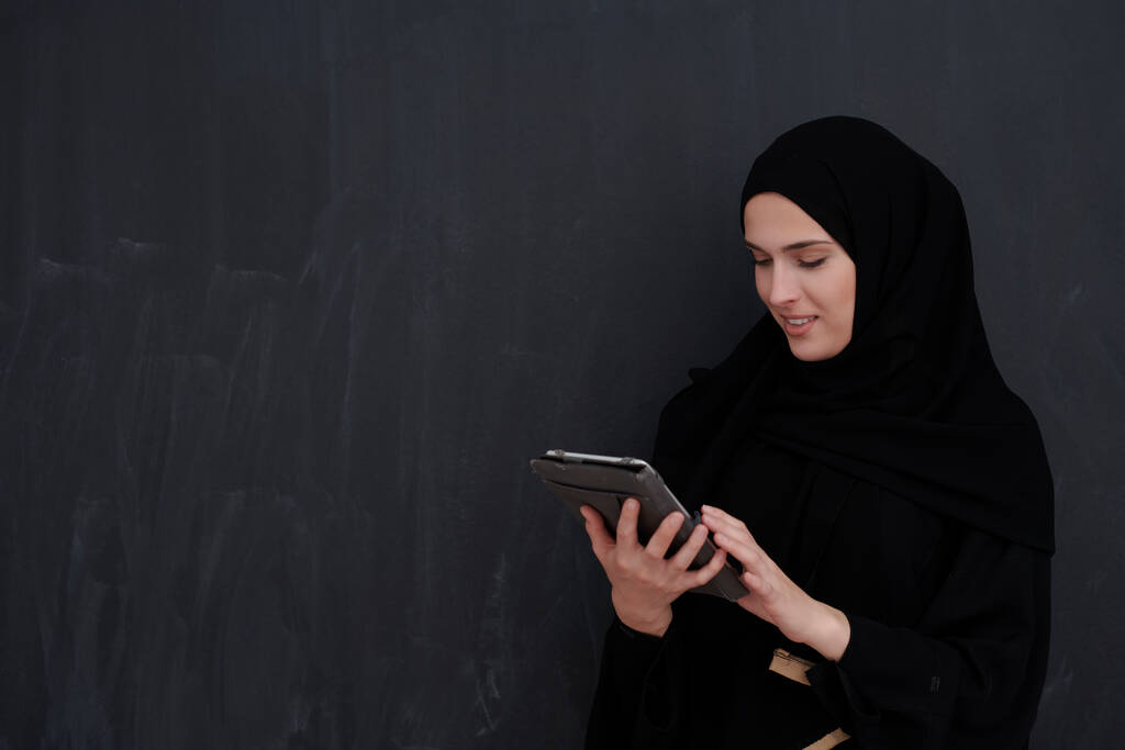 Giovane imprenditrice araba in abiti tradizionali o abaya e bicchieri con tablet davanti alla lavagna nera che rappresenta la moda islamica moderna e la tecnologia - Foto, immagini