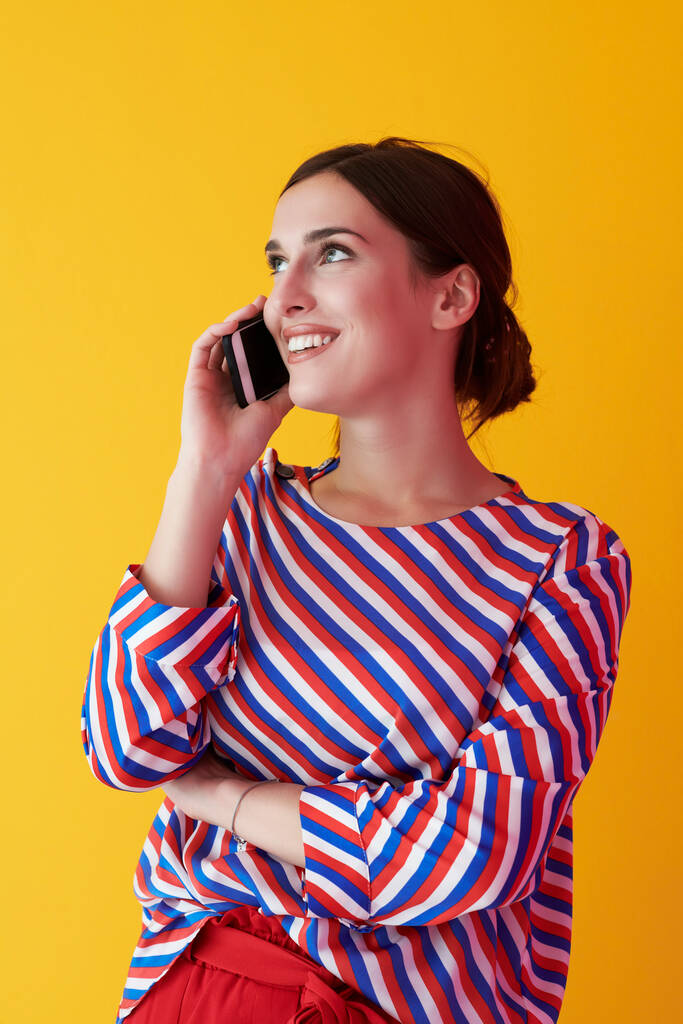 Portret młodej dziewczyny rozmawiającej przez telefon stojącej przed żółtym tłem. Kobieta modelka reprezentująca nowoczesną koncepcję mody i technologii - Zdjęcie, obraz