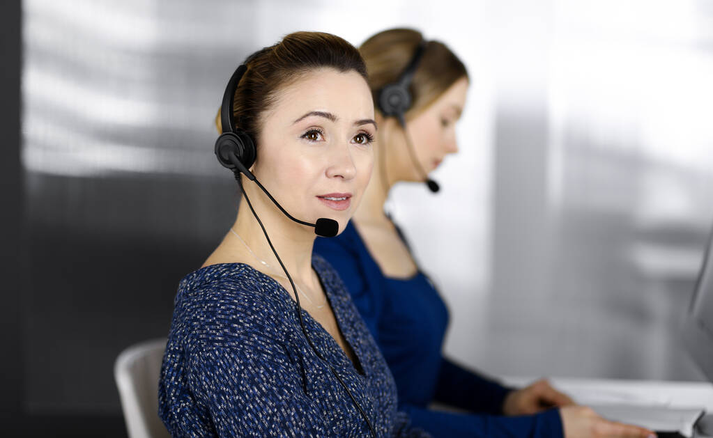 Zwei Geschäftsfrauen unterhalten sich per Headset mit den Kunden, während sie in einem modernen Büro am Schreibtisch sitzen. Diverse Menschen gruppieren sich in einem Callcenter. Telemarketing und Kundenservice - Foto, Bild