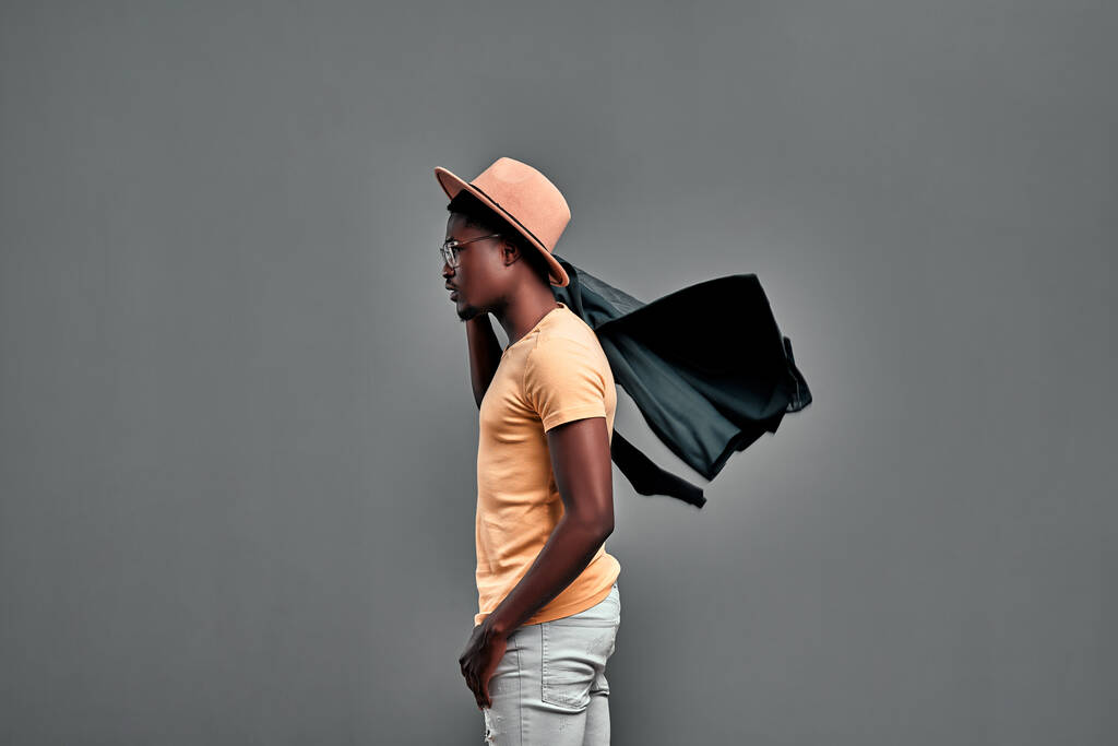 Een aantrekkelijke jonge Afrikaanse man die geïsoleerd over een grijze achtergrond loopt, met een leren jas op zijn schouder. Zijaanzicht. Kopieerruimte - Foto, afbeelding