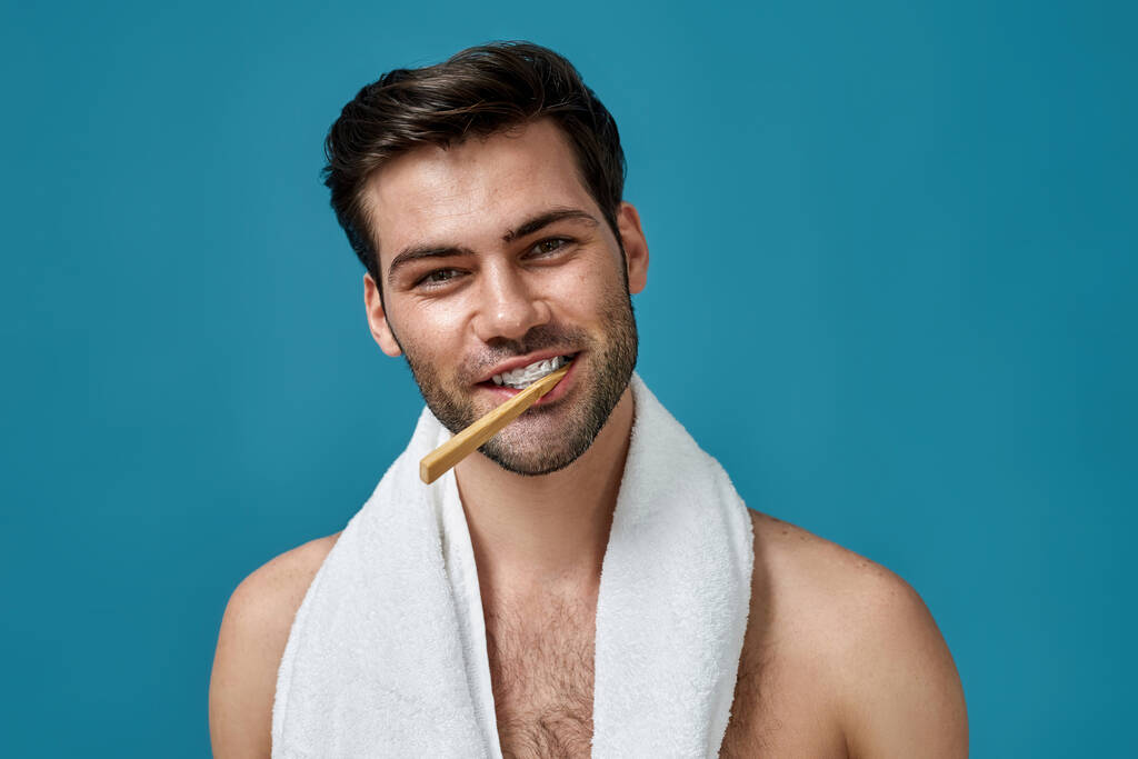 Porträt von sexy brünetten Kerl mit Handtuch um den Hals lächelt in die Kamera, während er mit Zahnbürste im Mund vor blauem Hintergrund posiert - Foto, Bild